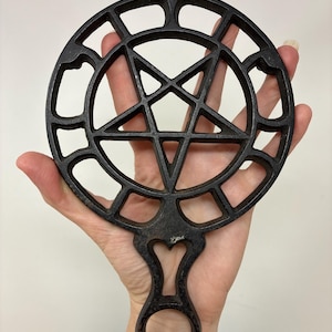 May include: A circular black cast iron trivet featuring a central pentagram design enclosed within a ring of rectangular cutouts. The trivet includes a handle with a heart-shaped cutout and a circular hole at the end for hanging purposes.