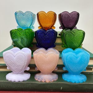 Puede incluir: Colección de nueve recipientes de vidrio en forma de corazón en varios colores, incluyendo azul, naranja, morado, verde y rosa. Cada recipiente tiene una superficie texturizada y se asienta sobre una base decorativa.