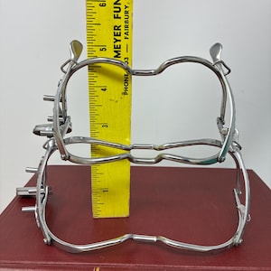 May include: Two silver-colored medical instruments are stacked on top of each other, with a yellow ruler placed between them. The ruler shows measurements in inches. The instruments are on a red book. The text "MEYER FUNERAL H" and "FAIRBURY, NEBRASKA" is visible on the ruler.