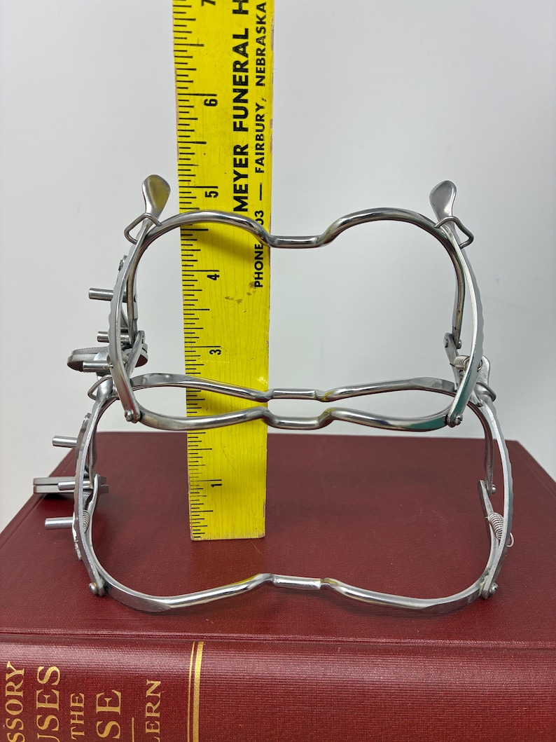 May include: Two silver-colored medical instruments are stacked on top of each other, with a yellow ruler placed between them. The ruler shows measurements in inches. The instruments are on a red book. The text "MEYER FUNERAL H" and "FAIRBURY, NEBRASKA" is visible on the ruler.