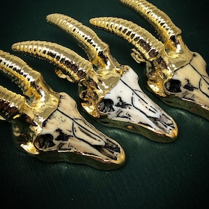 May include: Three gold-plated animal skull pendants with white bone details. The skulls have horns and are arranged in a row.