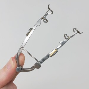 May include: A silver metal tool with two curved arms and a small knob. The arms have a gold-colored piece at the end.