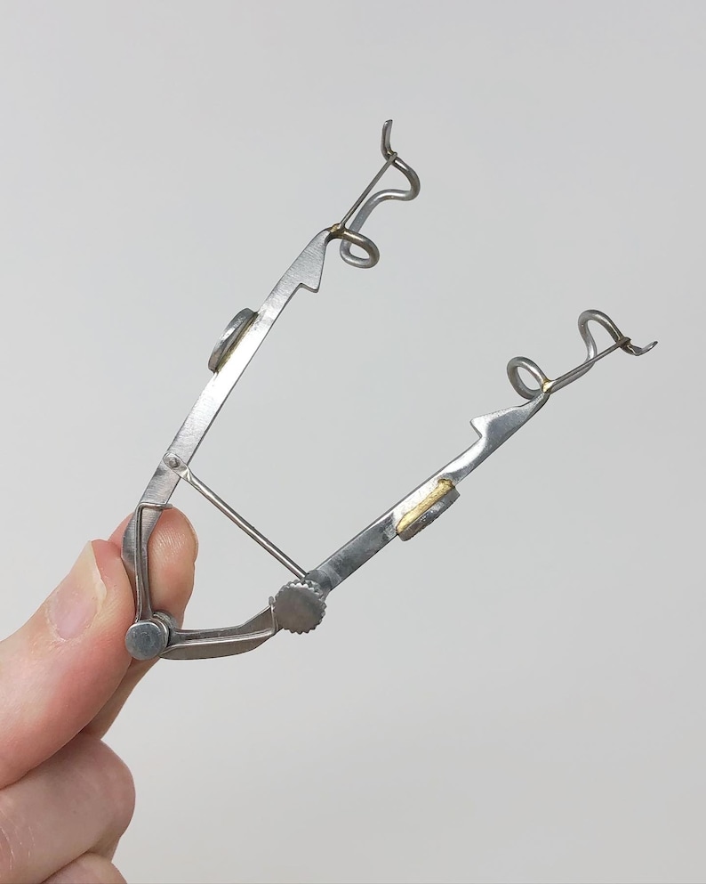 May include: A silver metal tool with two curved arms and a small knob. The arms have a gold-colored piece at the end.