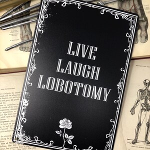 May include: Black metal sign with a white ornate border and a white rose. The sign reads "Live Laugh Lobotomy".