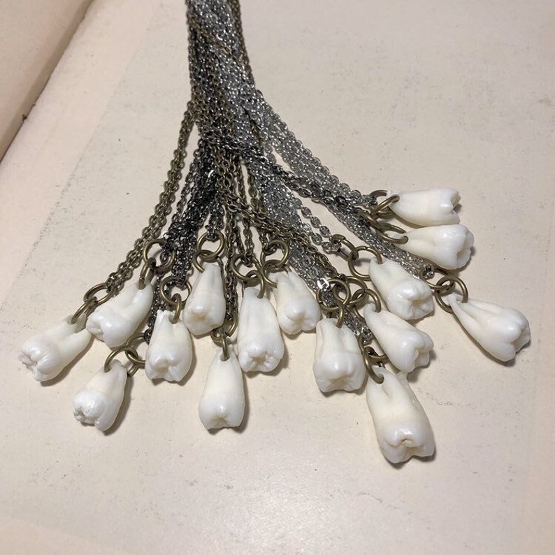 May include: A collection of white tooth charms hanging from silver and gold chains. The charms are all different sizes and shapes, and they are arranged in a fan shape.