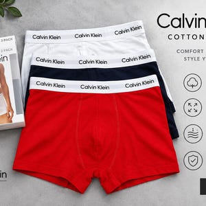 May include: A three-pack of Calvin Klein cotton stretch trunks in red, navy blue, and white. Each pair features a white elastic waistband with the Calvin Klein logo printed in black. The set includes a branded product box with a printed image.