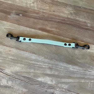 May include: A light green dog leash splitter with black metal clips and rivets. The splitter is made of a flexible material and is designed to connect to two dog collars. The leash is shown on a wooden surface.