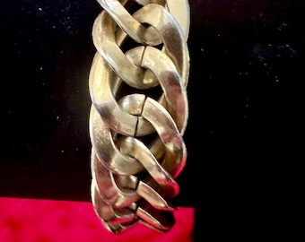 Puede incluir: Un brazalete de eslabones plateado. El brazalete presenta grandes eslabones ovalados entrelazados. Los eslabones están conectados en serie, formando un diseño audaz y grueso. El brazalete se muestra sobre un fondo negro.