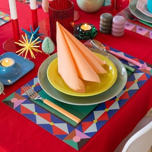 May include: A table setting featuring a peach-colored napkin folded on a yellow plate, which rests on a larger gray plate. A fork with a peach handle sits on a rectangular placemat with a geometric pattern of triangles and circles.