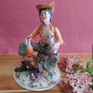 May include: A ceramic figurine of a seated man wearing an orange jacket, green pants, and a brown hat. He holds a bottle and a small dog. The base is decorated with floral patterns and a small barrel.