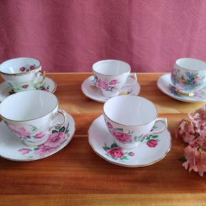 Puede incluir: Seis tazas de té y platillos de porcelana con diseños florales. Las tazas y los platillos tienen ribetes dorados y presentan rosas rosas y rojas, y otros motivos florales. Las tazas de té están dispuestas sobre una superficie de madera.