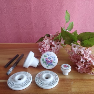 May include: A selection of white ceramic cabinet knobs and pulls with floral patterns. The knobs display blue, purple, and pink flowers. Two pulls and two backplates are also present. The items are arranged on a wooden surface with pink flowers.