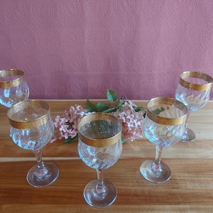May include: Five clear wine glasses with gold rims and etched designs are arranged on a wooden surface. A sprig of pink flowers is placed between the glasses. The background is a textured, mauve-colored wall.