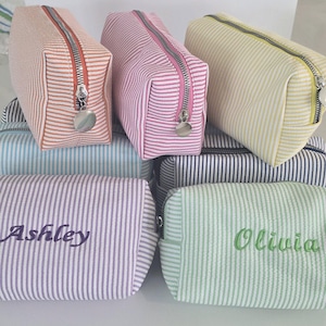 May include: A collection of rectangular fabric pouches featuring thin vertical stripes in various pastel colors including orange, pink, yellow, blue, purple, and green. Each pouch has a silver metal zipper and two are embroidered with names.