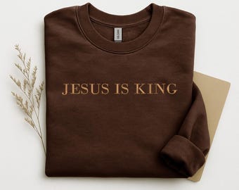 May include: A brown sweatshirt with the text "JESUS IS KING" in a gold color. The sweatshirt is folded with the sleeves cuffed. A sprig of dried flowers and a tan card are next to the sweatshirt.