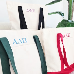 May include: Off-white canvas tote bags with embroidered Greek letters in purple, blue, and red. Each bag has a different color handle and trim: black, green, and red. The bags are displayed in a well-lit setting.