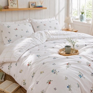 May include: White floral bedding set with a duvet cover and two pillowcases. The bedding features a pattern of small blue, red, and yellow flowers. A woven tray with a mug and vase sits on the bed. The room has a bright, airy feel.