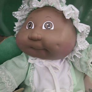 May include: A vintage baby doll with dark skin, wearing a mint green dress and a white lace-trimmed bonnet. The doll has large, expressive eyes and rosy cheeks, with a soft, rounded face. The doll's outfit includes a white bib with a bow.