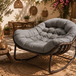 May include: A rounded rocking chair featuring a dark brown metal frame with woven detailing and a thick, tufted grey fabric cushion. The chair has a wide, curved seat design and sits on two curved rockers, providing a deep, comfortable seating area.