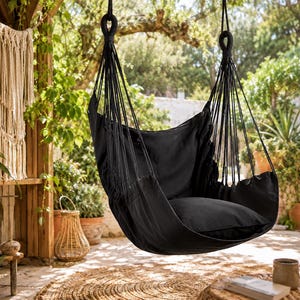 Puede incluir: Una silla colgante negra con un cojín suave, suspendida de una rama de árbol. La silla está hecha de una tela oscura y tiene múltiples cuerdas de soporte. La escena está ambientada al aire libre con luz natural y vegetación.