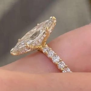 Puede incluir: Anillo de compromiso con diamante talla marquesa y banda dorada. El anillo presenta un diamante grande y transparente en forma de marquesa en el centro, rodeado de diamantes más pequeños. La banda también está adornada con diamantes más pequeños, creando un efecto brillante.