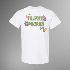 Puede incluir: Camiseta blanca con el texto "ALPHA MICRON PI" en verde, decorada con flores coloridas. El diseño tiene una estética retro y groovy. La camiseta es de estilo cuello redondo clásico.