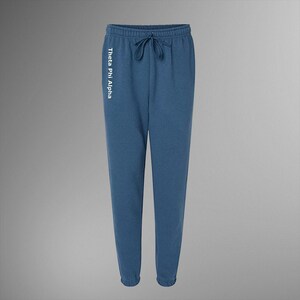 Puede incluir: Pantalones de chándal azules con cintura con cordón y puños elásticos. El texto "Theta Phi Alpha" está impreso verticalmente en la pierna izquierda en blanco. Los pantalones están hechos de un material suave.