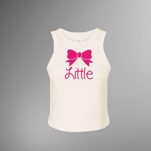 Puede incluir: Camiseta sin mangas color crema con un gráfico de lazo rosa brillante y la palabra "Little" en una escritura rosa a juego. La camiseta sin mangas tiene un cuello redondo y está hecha de un material suave y ligero.