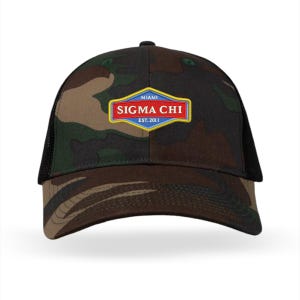 Puede incluir: Una gorra de béisbol de camuflaje con la parte trasera de malla negra. La parte delantera de la gorra presenta un parche con el texto "MIAMI SIGMA CHI EST. 2011" en rojo, azul y amarillo.