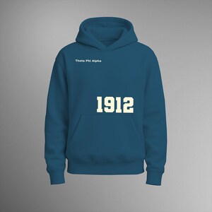 Op de afbeelding: Een tealkleurige hoodie met een voorzak en een capuchon. De tekst "Theta Phi Alpha" is boven de voorzak gedrukt en het nummer "1912" staat eronder in crème.