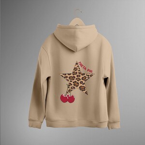 Puede incluir: Sudadera con capucha de color beige con un diseño de estrella con estampado de leopardo y el texto "THETA PHI" en rojo. Debajo de la estrella hay dos cerezas rojas. La sudadera con capucha está colgada de una percha.