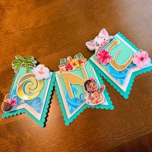 May include: A banner with the letters "ONE" in gold, featuring tropical flowers, palm trees, and images of characters from a popular animated film. The banner has a scalloped edge and is in shades of teal, white, and gold.