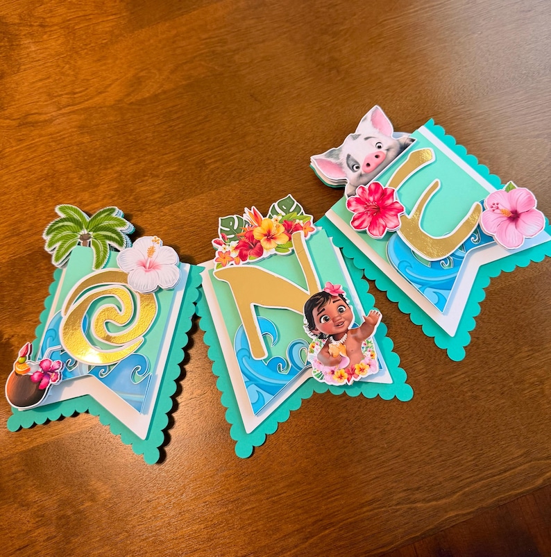 May include: A banner with the letters "ONE" in gold, featuring tropical flowers, palm trees, and images of characters from a popular animated film. The banner has a scalloped edge and is in shades of teal, white, and gold.