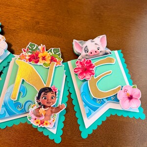 May include: A banner with the letters "NE" in gold, featuring illustrations of a baby girl and a pig. The banner has a teal scalloped edge, with tropical flowers and wave designs. The background is a wooden surface.