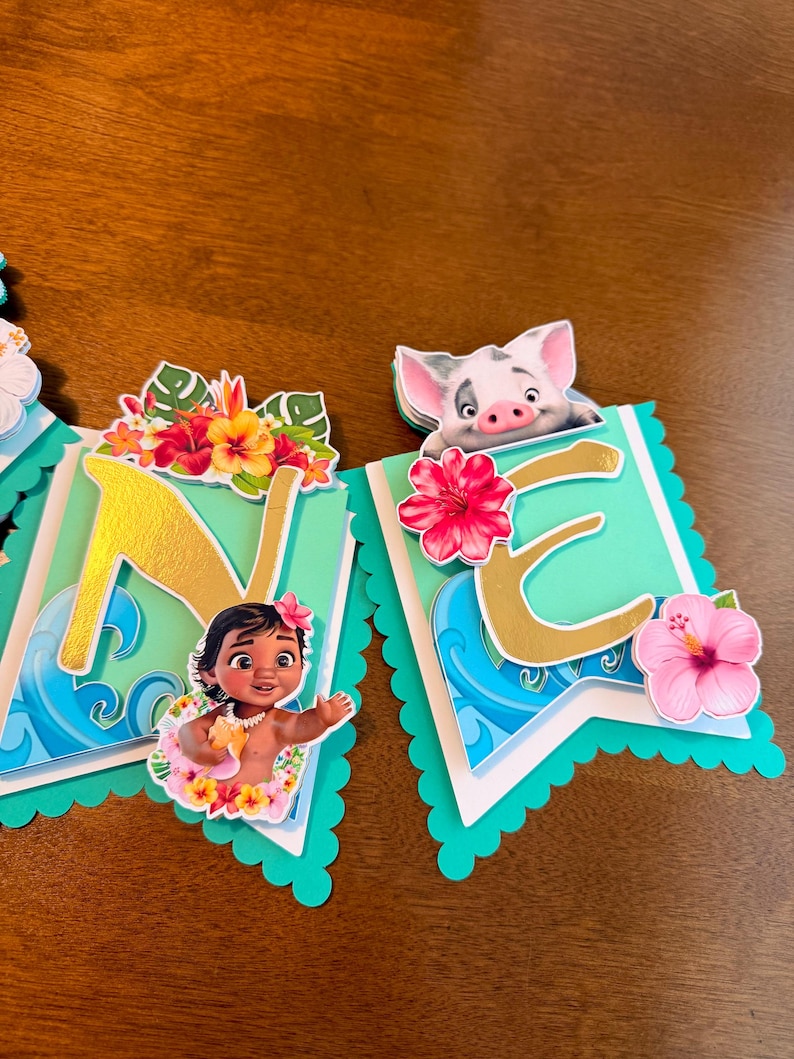 May include: A banner with the letters "NE" in gold, featuring illustrations of a baby girl and a pig. The banner has a teal scalloped edge, with tropical flowers and wave designs. The background is a wooden surface.