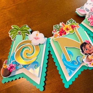 May include: A banner with a tropical theme, featuring cut-out letters and images. The banner has a teal scalloped edge and includes images of flowers, palm trees, and characters. The letters are gold and the background has a wave design.
