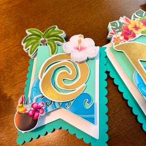 May include: A banner with a tropical theme, featuring a teal scalloped edge. The banner includes a gold design, blue waves, a palm tree, a white hibiscus flower, and a coconut drink with a flower. The banner is layered with white and teal paper.