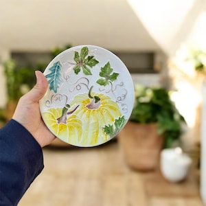 May include: A white ceramic plate with a hand-painted design of two yellow pumpkins and green leaves. The plate has a scalloped edge and is held in a person's hand. The plate is a decorative item.
