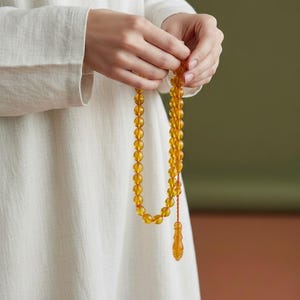 May include: A string of translucent, honey-colored amber prayer beads featuring round, polished spheres. The beads are connected by a thin, orange-toned cord and terminate in a single, elongated, teardrop-shaped pendant piece at the bottom.