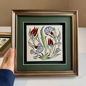 May include: Framed ceramic tile featuring a floral design with red tulips and blue flowers on a white background. The tile is framed with a gold-colored frame and a green matting.