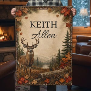 May include: A rectangular woven blanket featuring a deer standing in a forest landscape with mountains and a lake. The design includes a border of autumn leaves and pine needles. The text "KEITH Allen" is printed in the center in black font.