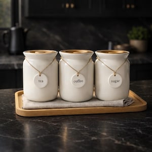 May include: Three cream-coloured ceramic storage jars with gold lids and labels reading "tea", "coffee", and "sugar". Each jar hangs from a gold chain. The jars sit on a wooden tray with a beige cloth.