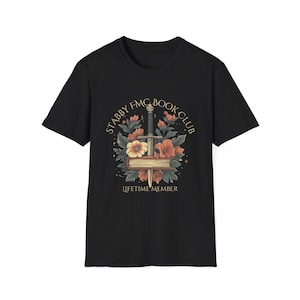 May include: Black t-shirt with a gold graphic of a book with a sword through it and flowers. The text "Stabby FMC Book Club" and "Lifetime Member" are also on the shirt.
