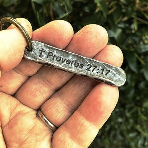 May include: A silver-colored metal keychain with the text "Proverbs 27:17" engraved on it. The keychain has a hammered texture and a bronze-colored ring. The background is blurred green foliage.
