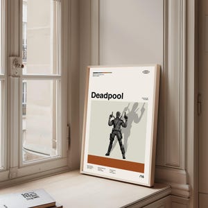 Puede incluir: Un p&oacute;ster enmarcado con la palabra "Deadpool" en negro. El p&oacute;ster muestra una ilustraci&oacute;n en escala de grises del personaje de Deadpool sosteniendo dos armas, con una sombra detr&aacute;s. El fondo es de color beige claro.