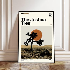 Op de afbeelding: Een ingelijste poster met de albumhoes van U2's "The Joshua Tree". Het ontwerp toont een zwart silhouet van een Joshua-boom tegen een woestijnlandschap met een oranje zon. De titel en het jaar 1987 zijn afgebeeld.