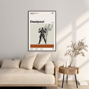 Puede incluir: Un p&oacute;ster enmarcado con la palabra "Deadpool" en negro. El p&oacute;ster muestra una figura con un traje negro con armas levantadas, proyectando una sombra. El fondo es beige y marr&oacute;n. El p&oacute;ster se muestra en una pared blanca.