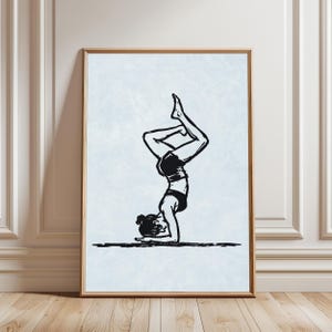 May include: A rectangular wall art print featuring a black ink illustration of a person performing a forearm stand yoga pose. The figure is depicted in a high-contrast, woodcut style against a textured, pale light blue background within a wooden frame.