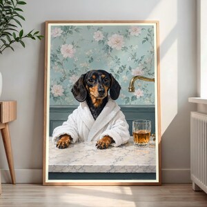 May include: A framed wall art print featuring a black and tan dachshund wearing a white plush bathrobe. The dog rests its paws on a marble countertop next to a glass of amber liquid. The background shows floral wallpaper and a brass faucet.
