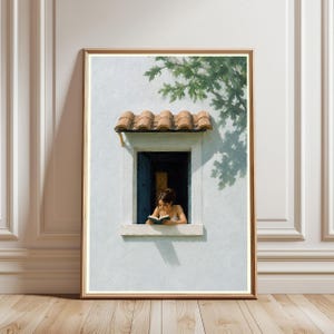 May include: A framed wall art print depicting a person reading a book while leaning on the sill of an open window. The window features dark blue shutters and a terracotta tiled roof, set against a light gray wall with green tree shadows.